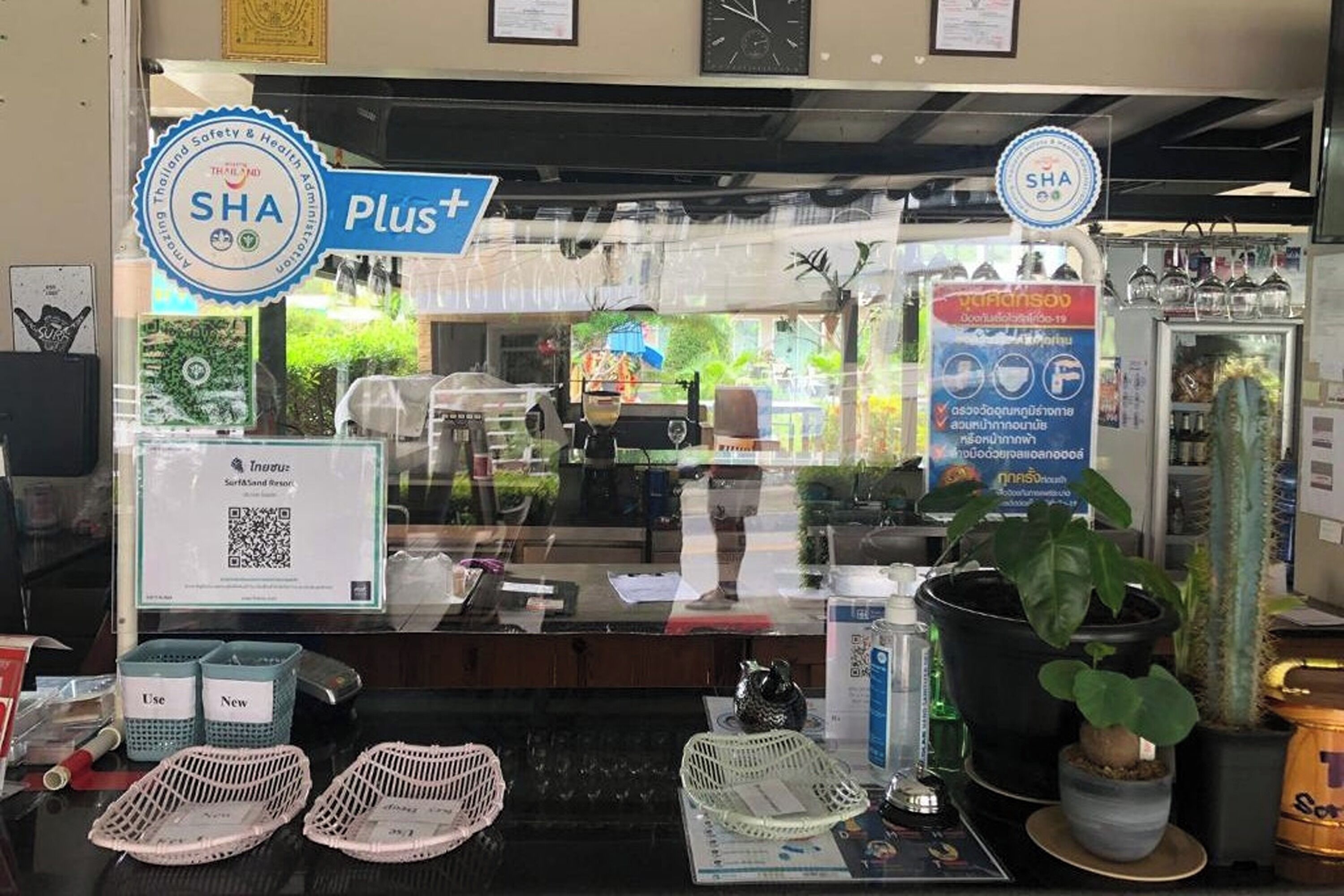 Fachada con un cartel que indica 'SHA Plus+' y un emblema de 'Thailand Safety & Health Administration'. Hay cestas con las etiquetas 'Usar' y 'Nuevas', un código QR y una exhibición de cestas tejidas.