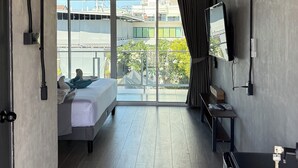 Quarto de hotel moderno com cama, televisão, escrivaninha e varanda com vista para edifícios e vegetação.