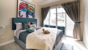 Quarto com cama, manta branca e fofa, obra de arte com elefante emoldurada e vista pela janela grande.
