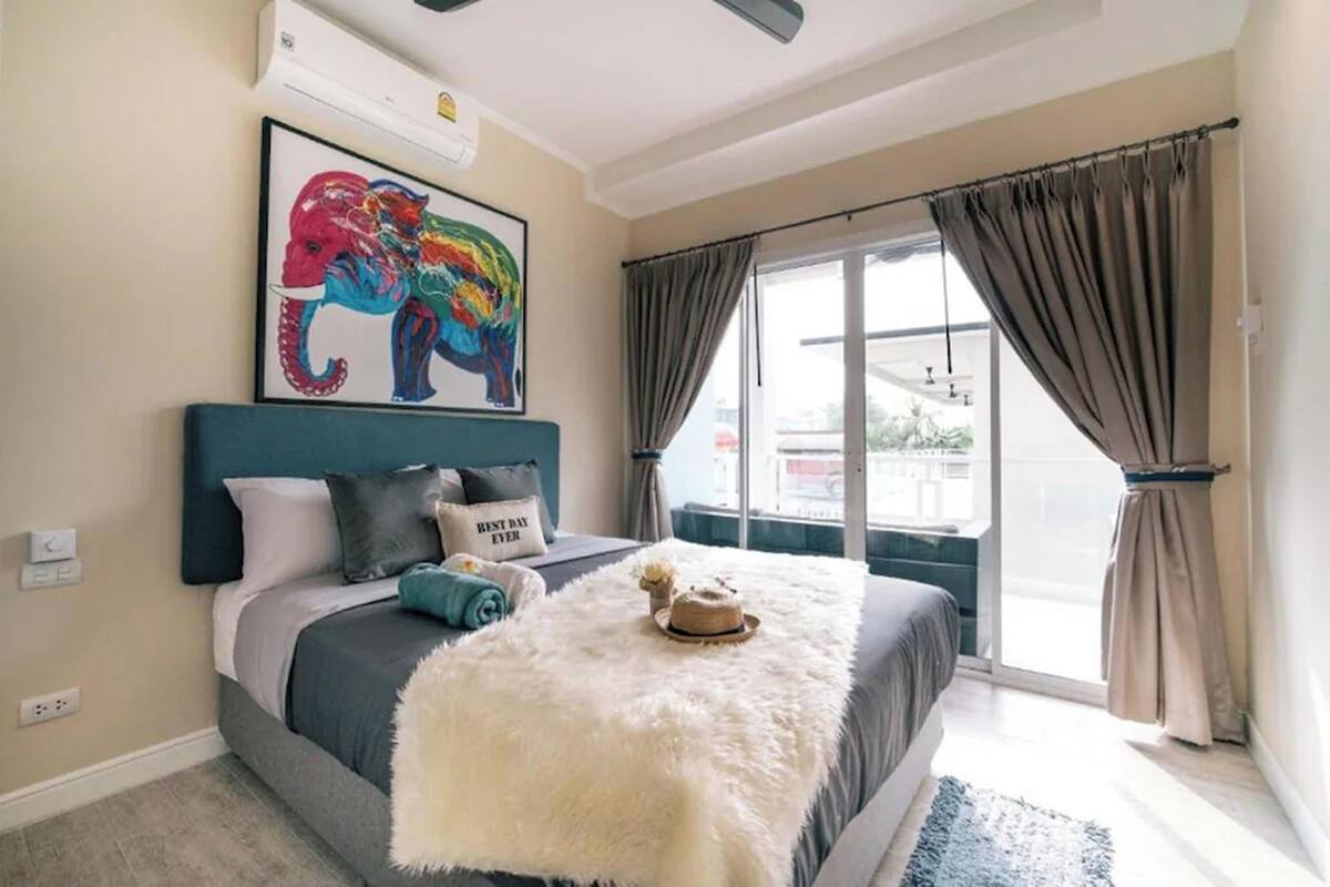 Une chambre à coucher avec un lit, un plaid blanc moelleux, un tableau représentant un éléphant encadré et une vue dégagée sur l’extérieur grâce à une grande fenêtre.