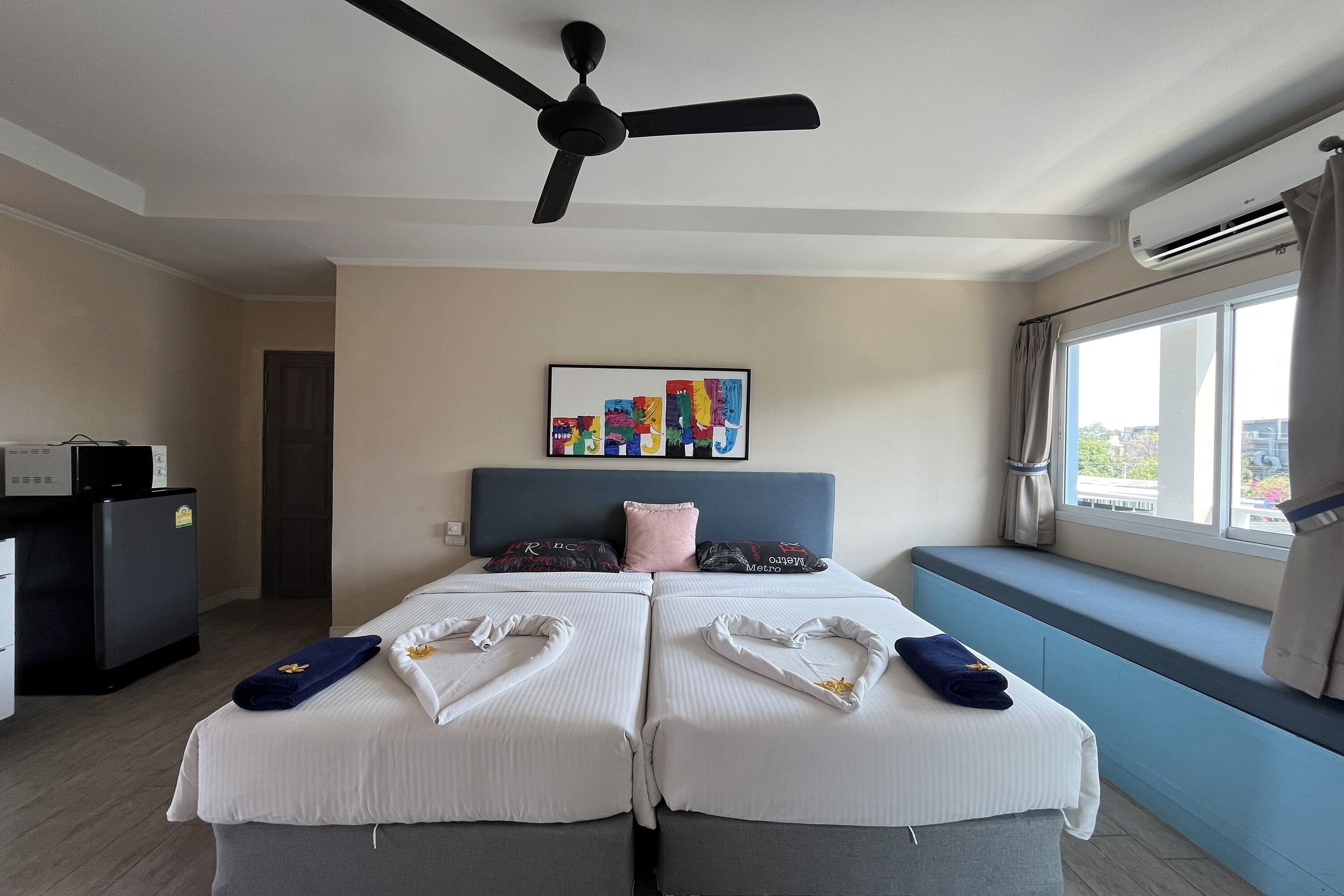 A hotel room with a bed, a ceiling fan, an air conditioning unit, a window with curtains, and a painting on the wall.