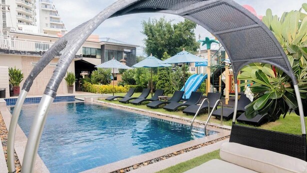 Piscina com espreguiçadeiras e guarda-sóis, cercada por vegetação e edifícios.
