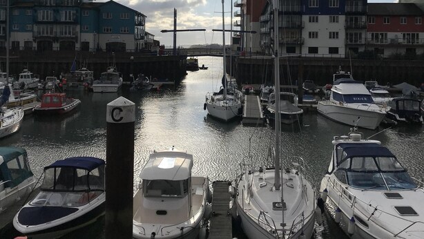A marina with boats docked, a wooden dock, and buildings in the background.