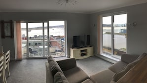A living room with a sofa, a TV on a stand, and a view of the sea through large windows.