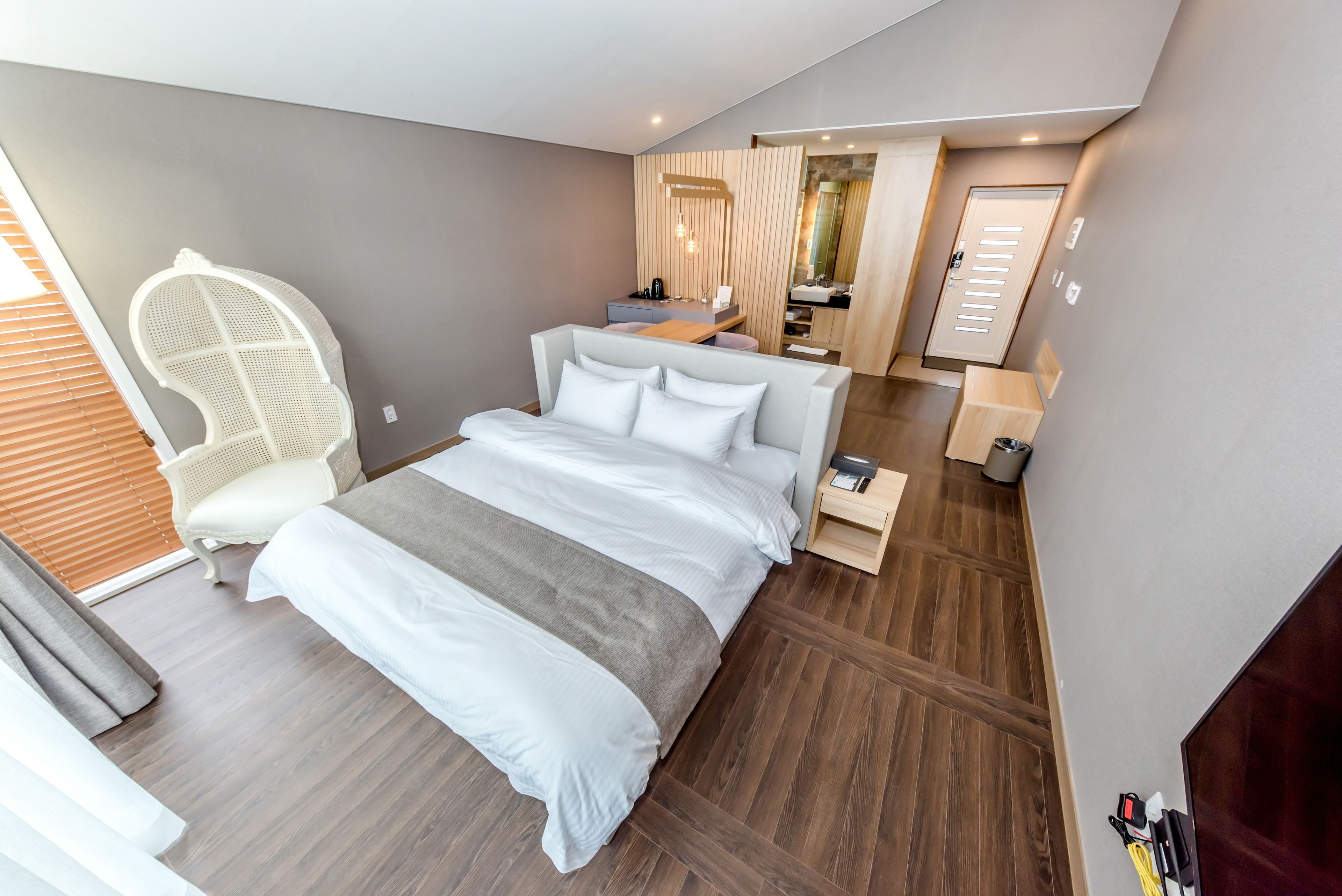 A modern hotel room with a large bed, a seating area with a chair, a small desk, and a bathroom visible through an open door.
