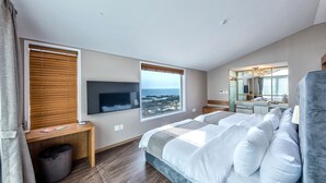 A modern bedroom with a large bed, a TV mounted on the wall, and a view of the ocean through the windows.