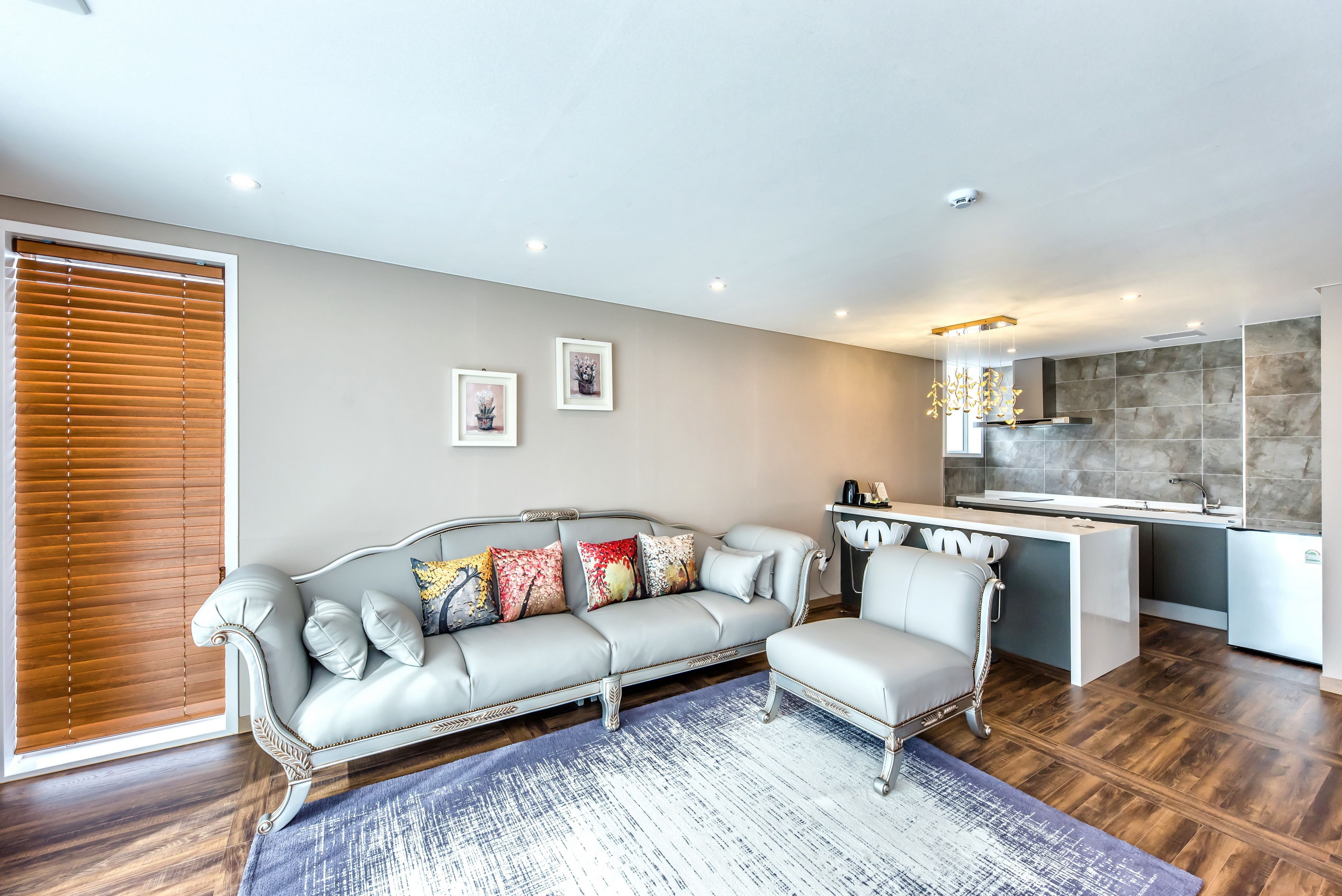 A modern living room with a grey sofa, a small ottoman, and a kitchen area with a white countertop and grey tiles.