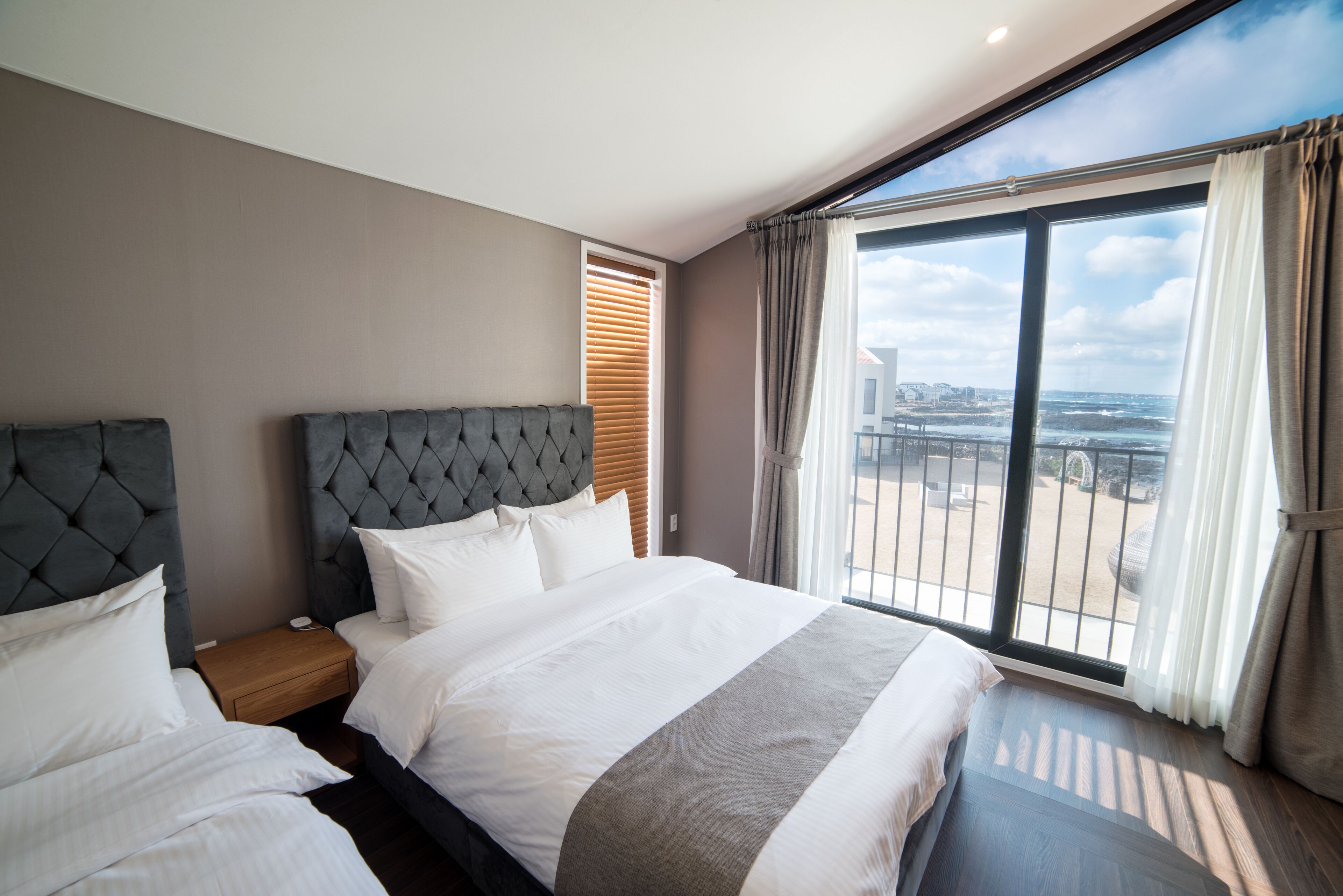 A modern hotel room with a large bed, a nightstand, and a view of the city through large windows.