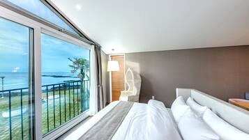 A modern hotel room with a large window, a bed with white linens, a wooden headboard, and a view of the sea.