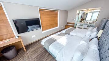 Quarto moderno com uma cama grande, televisão montada na parede, janela com persianas, toucador com espelho e lixeira.