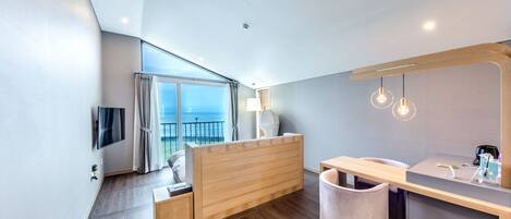 A modern hotel room with a wooden bed, a desk, a chair, a trash bin, a TV, and a large window with a view of the sea.
