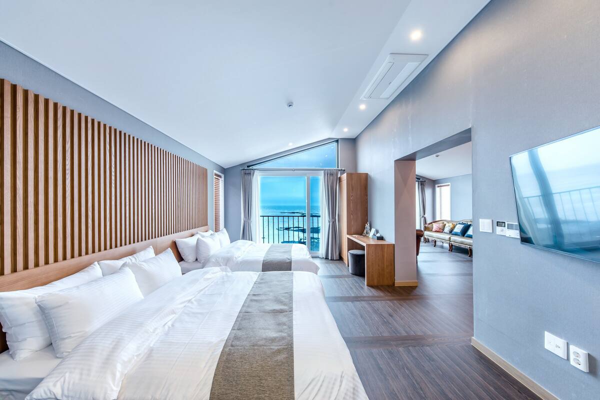 Ein modernes Hotelzimmer mit einem großen Bett, einem Lattenrost-Kopfteil, einer Sitzecke mit Sofa und Sesseln sowie einem großen Fenster mit Blick aufs Meer.