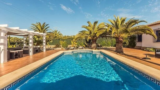 A rectangular swimming pool with clear blue water, surrounded by palm trees and a pergola. - Fully Air Conditioned Villa With Heated Pool Set In Large Landscaped Gardens (Alicante)
