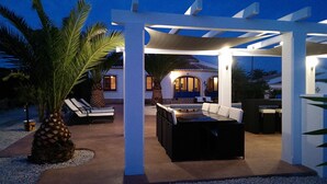 An outdoor patio area with a covered seating arrangement, a dining table, and a large potted plant. - Fully Air Conditioned Villa With Heated Pool Set In Large Landscaped Gardens (Alicante)