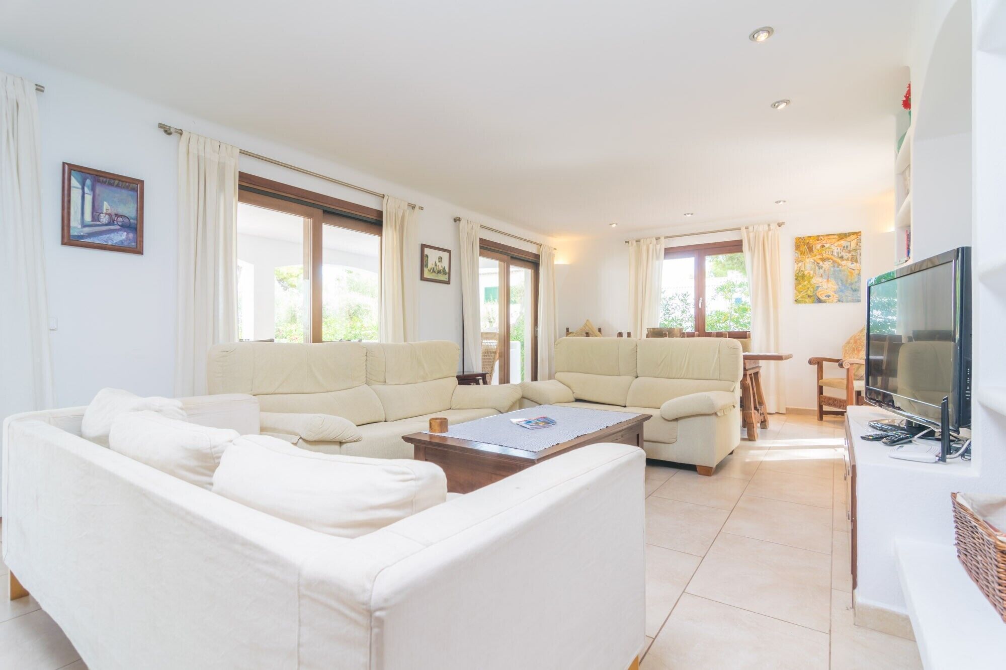 Ein geräumiges Wohnzimmer mit einer großen, beigefarbenen Eckcouch, einem Couchtisch und einem Fernseher. Große Fenster mit Vorhängen, ein Gemälde an der Wand und ein kleiner Tisch mit einem Stuhl sind ebenfalls vorhanden.