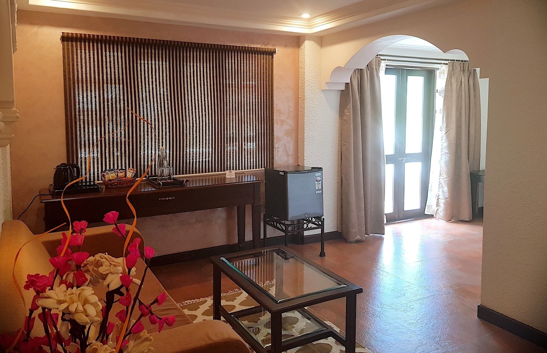 Une chambre d’hôtel avec un canapé, une table basse en verre, une plante décorative, un minibar et une vue sur l’extérieur par une porte munie de rideaux.