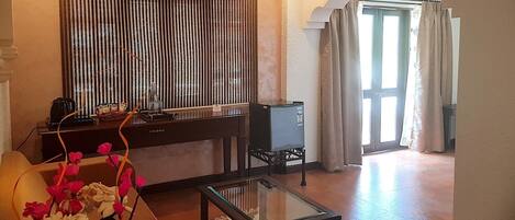 Une chambre d’hôtel avec un canapé, une table basse en verre, une plante décorative, un minibar et une vue sur l’extérieur par une porte munie de rideaux.