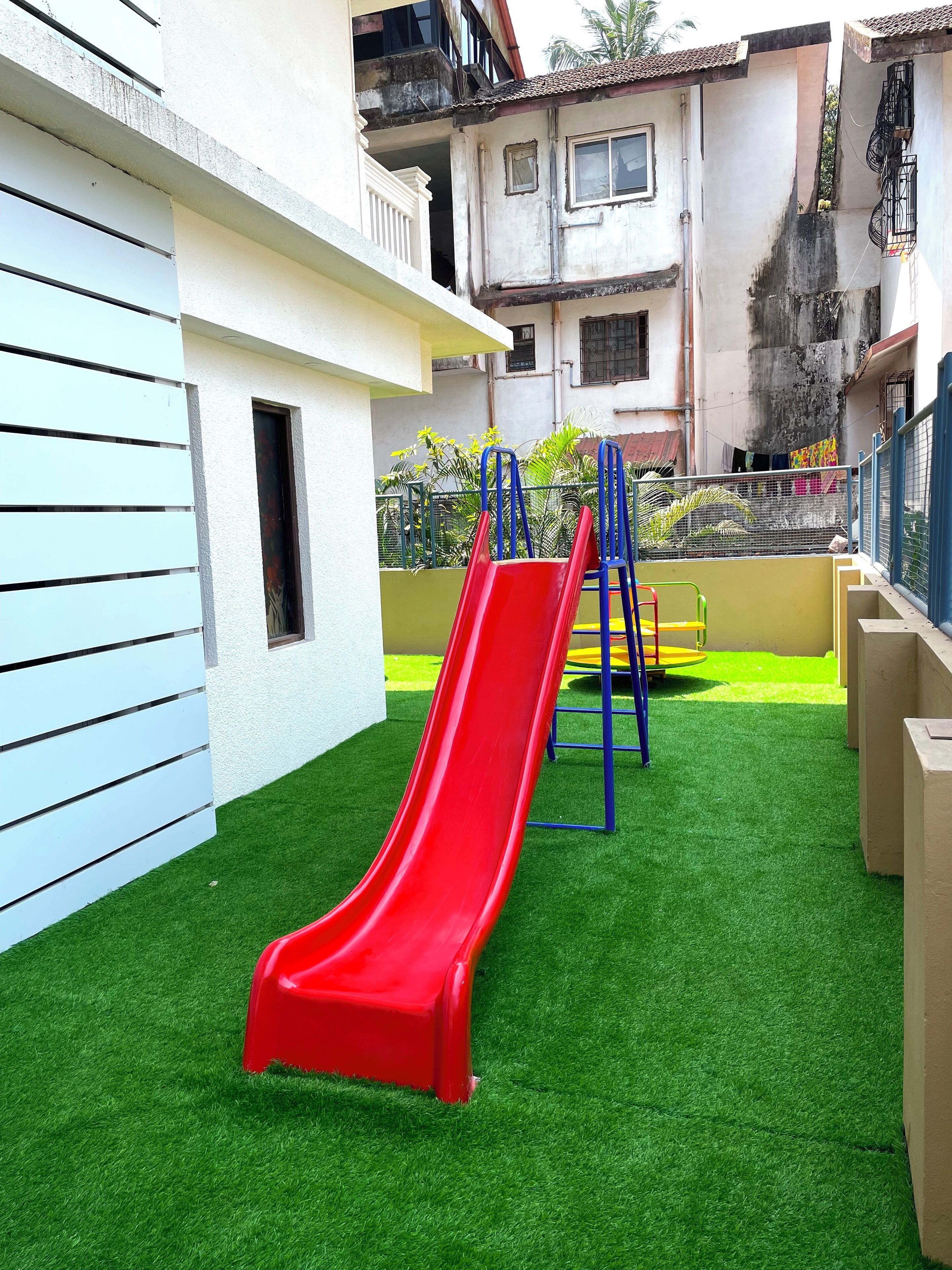 Une aire de jeux avec un toboggan rouge et des structures d’escalade bleues, installée sur une pelouse artificielle et entourée de bâtiments.