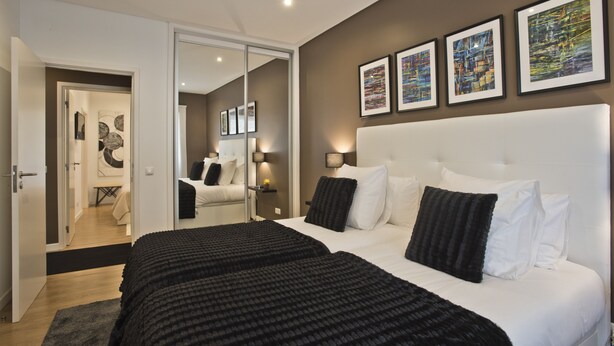 A modern bedroom with a large bed, a white headboard, and a mirror. There are framed pictures on the wall and a door leading to another room. - A modern apartment in Cascais Center (Cascais)