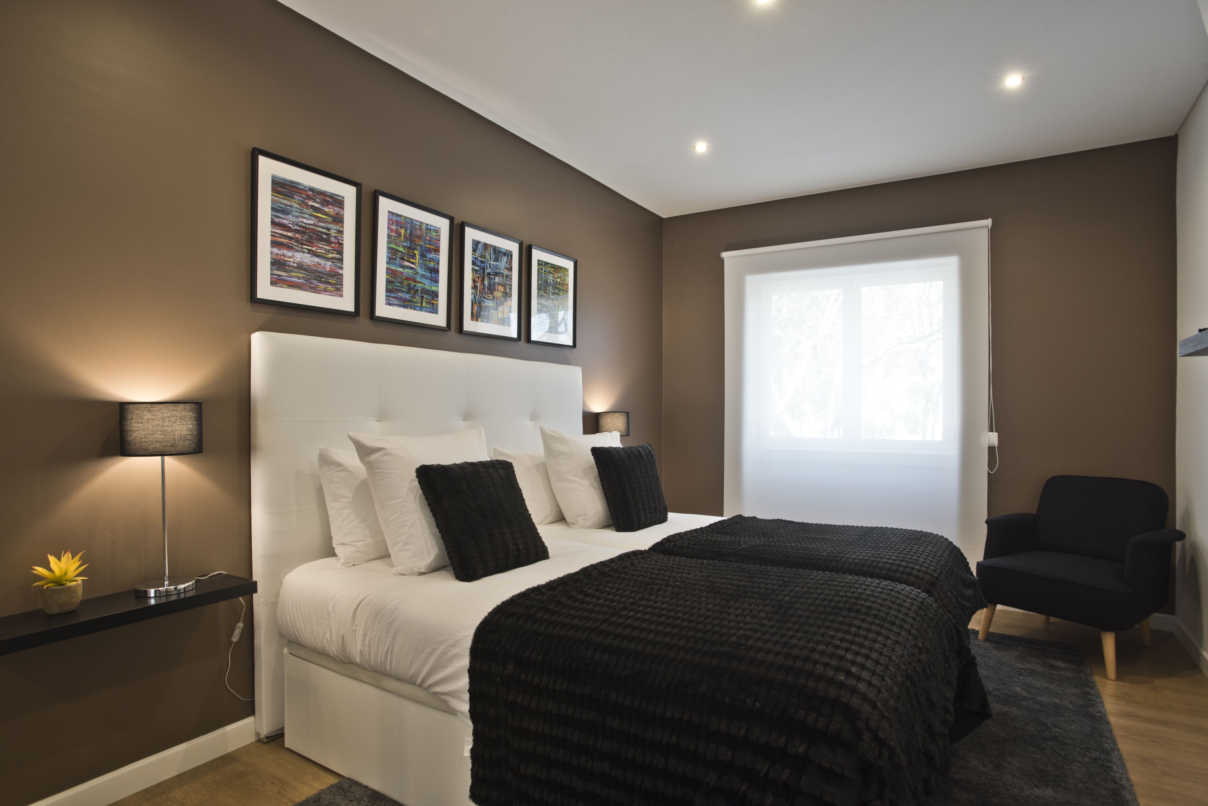 Ein modernes Schlafzimmer mit einem großen Bett, einem Nachttisch, einer Lampe und einem Sessel. An der Wand hängen drei gerahmte Bilder.