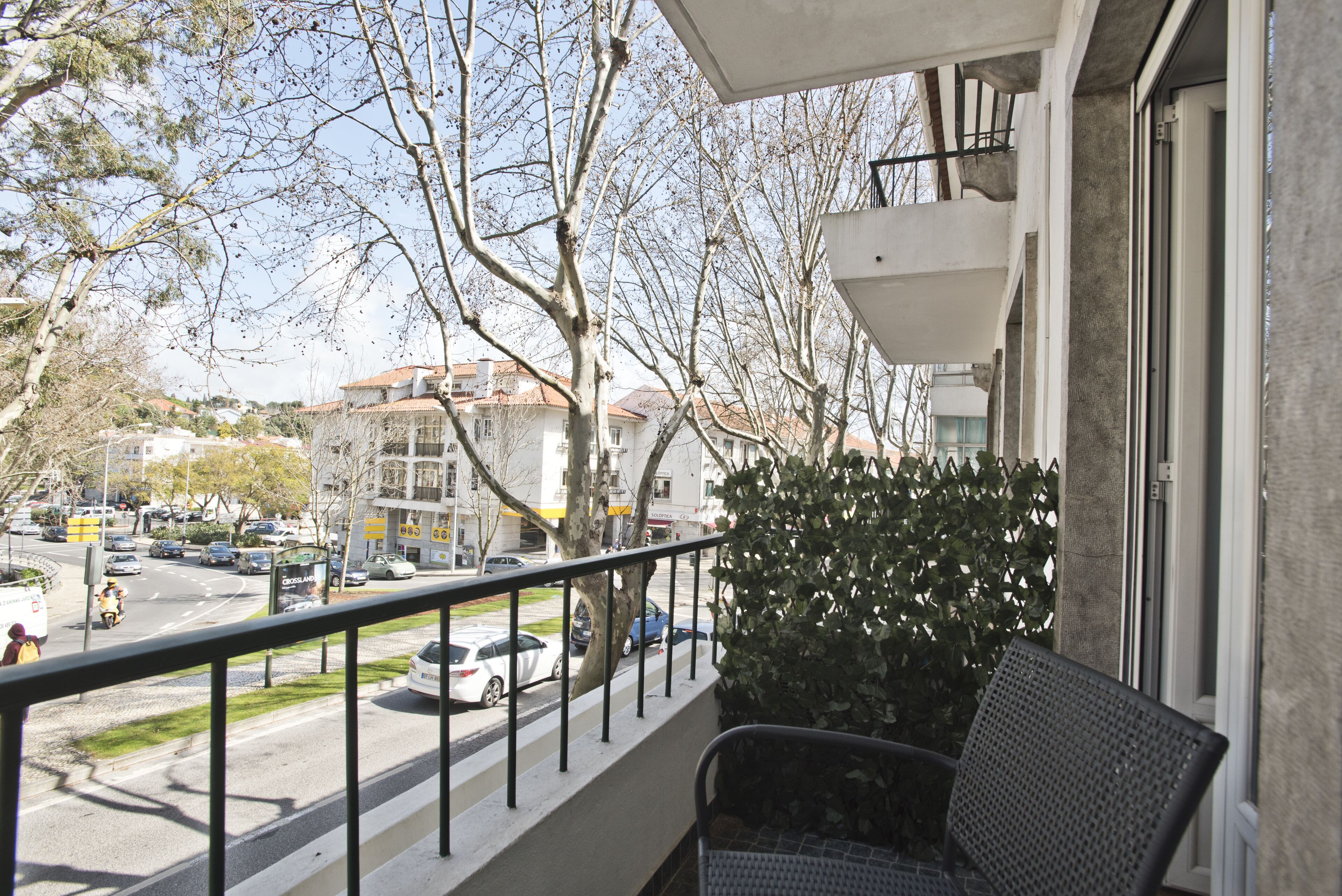 A balcony with a view of a street, a hedge, and a building with a balcony.