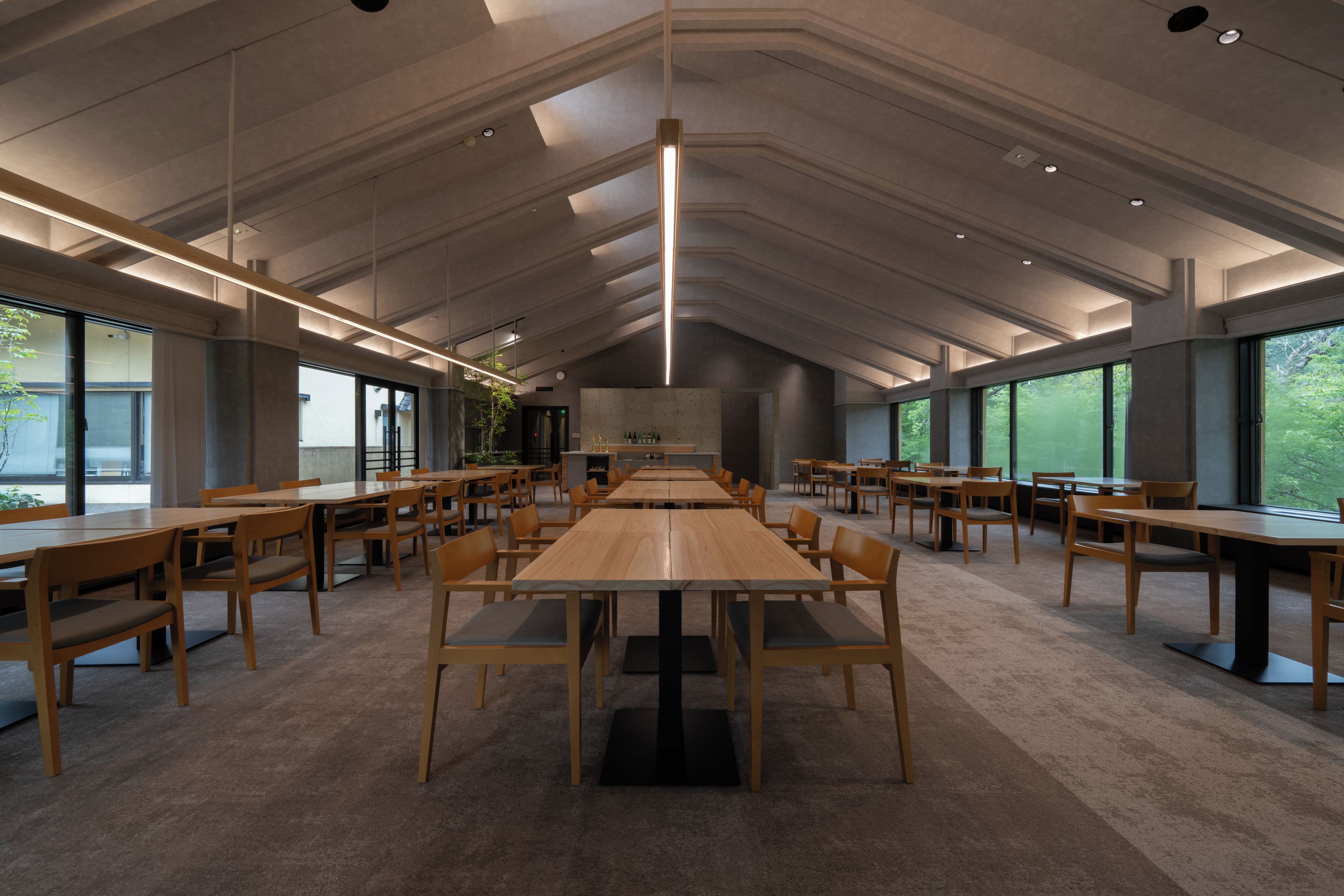 A spacious indoor area with multiple wooden tables and chairs, large windows, and a high ceiling with recessed lighting.