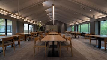 A spacious indoor area with multiple wooden tables and chairs, large windows, and a high ceiling with recessed lighting.