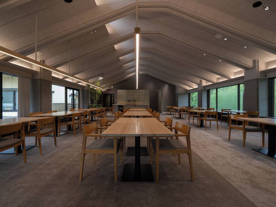 Un espacio interior amplio con varias mesas y sillas de madera, ventanas grandes y un techo alto con iluminación empotrada.