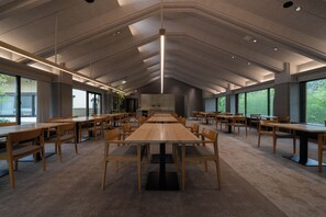 Un vaste espace intérieur doté de nombreuses tables et chaises en bois, de grandes fenêtres et d’un plafond haut avec des luminaires encastrés.