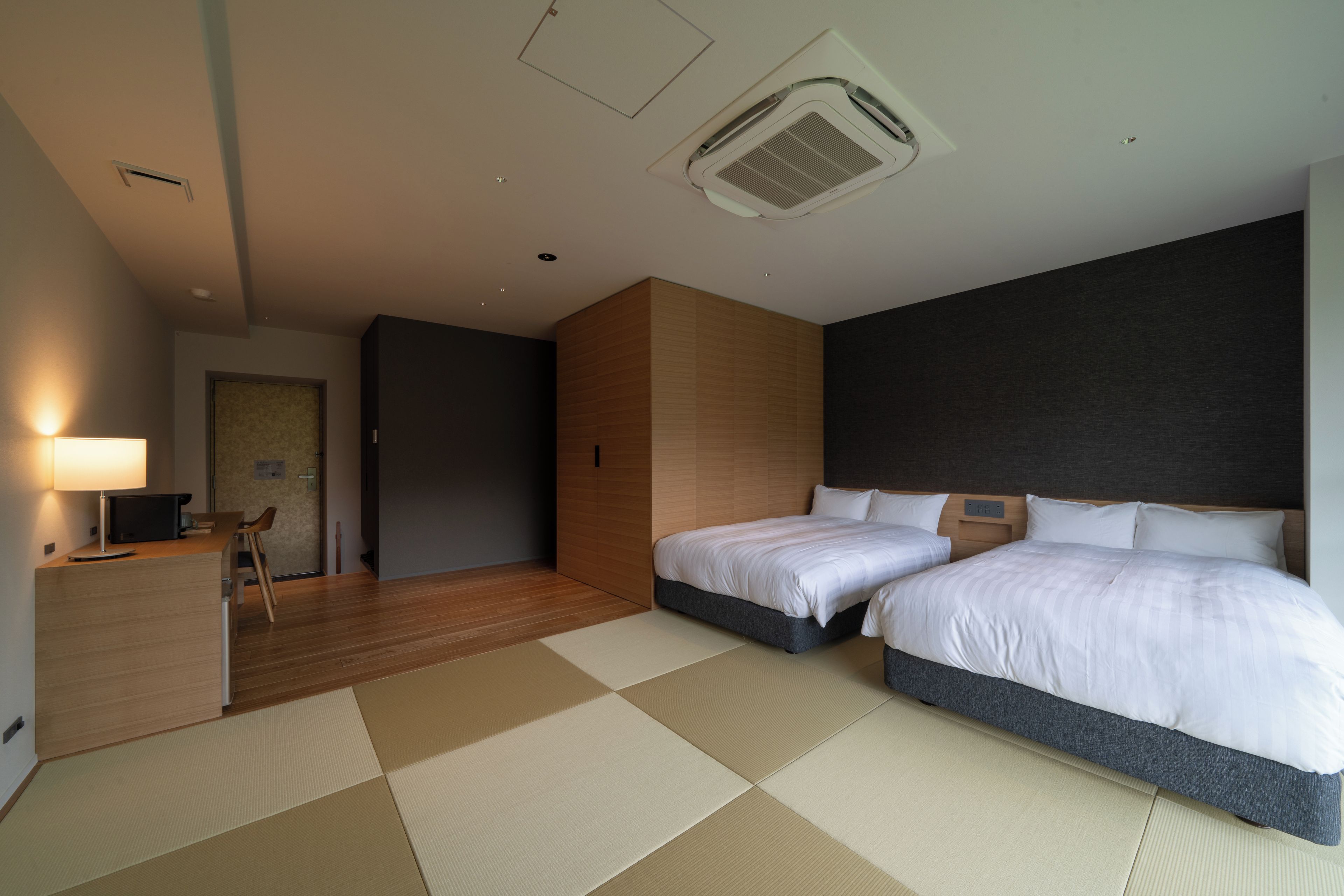 A hotel room with two beds, a desk with a chair, a lamp, and a large window.