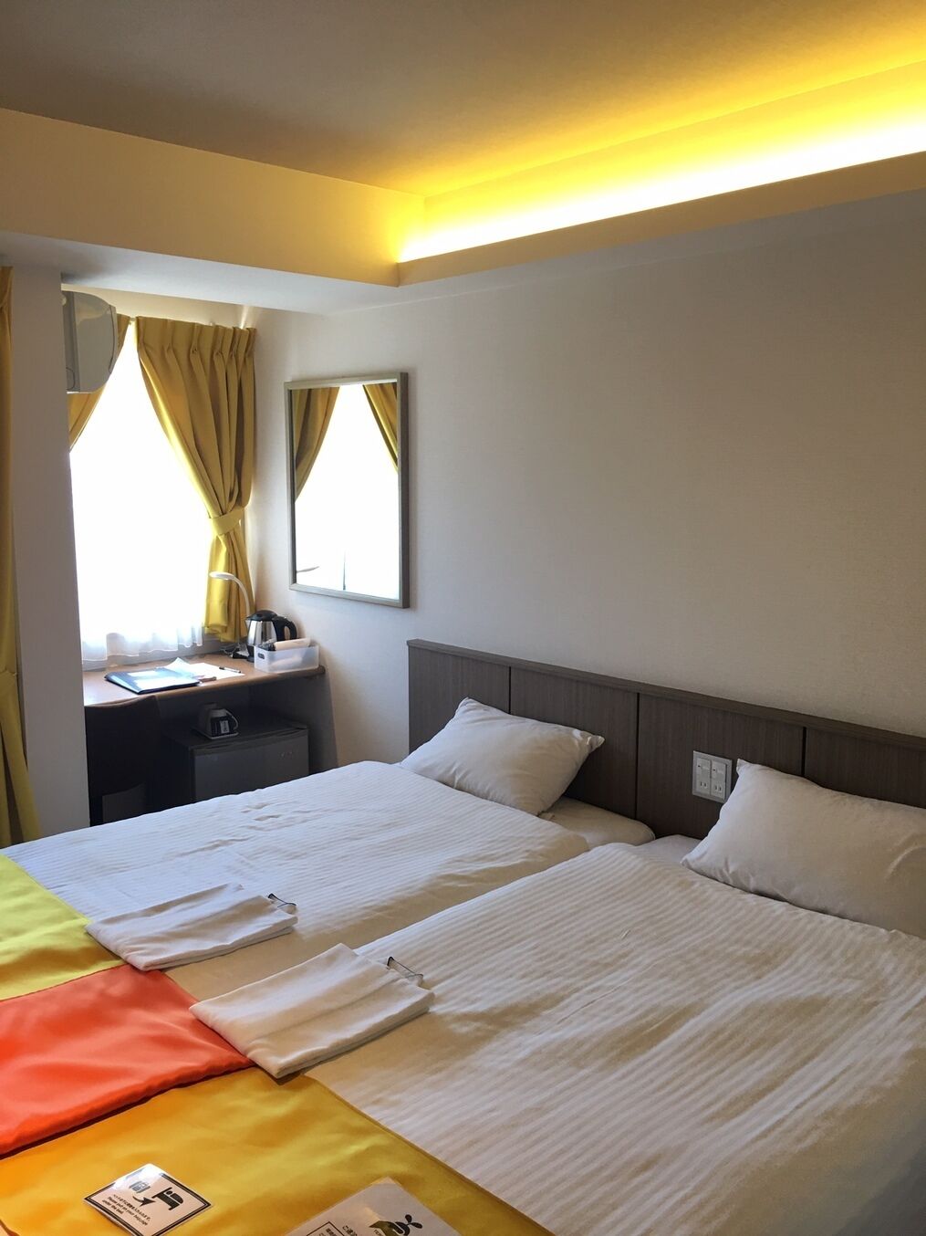 A hotel room with two beds, a desk with a chair, a mirror, and a window with curtains.