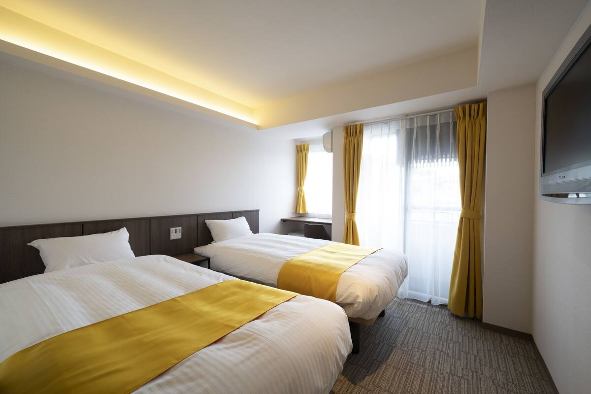 A hotel room with two beds, a flat-screen TV, a desk, and a window with curtains.