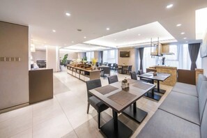 Un comedor moderno de hotel con varias mesas y sillas, una zona de cocina al fondo y amplias ventanas que ofrecen luz natural.