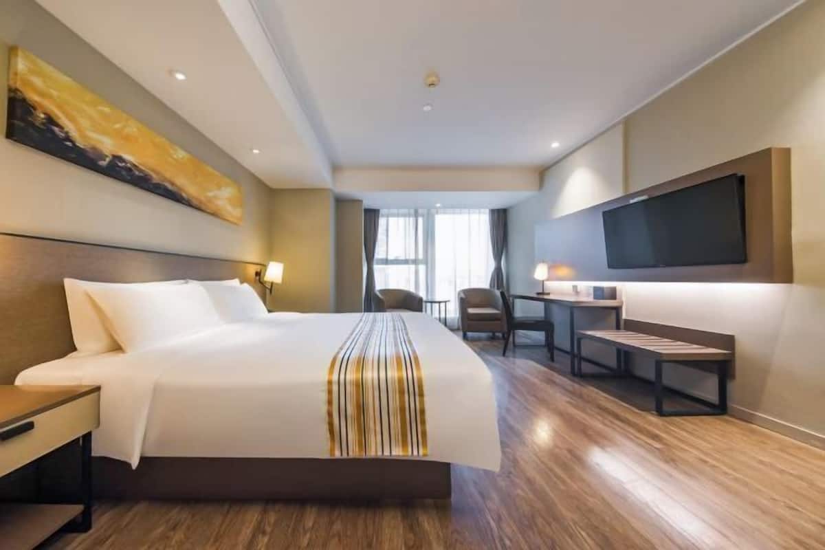 A hotel room with a large bed, a desk with a chair, a television mounted on the wall, and a window with curtains.