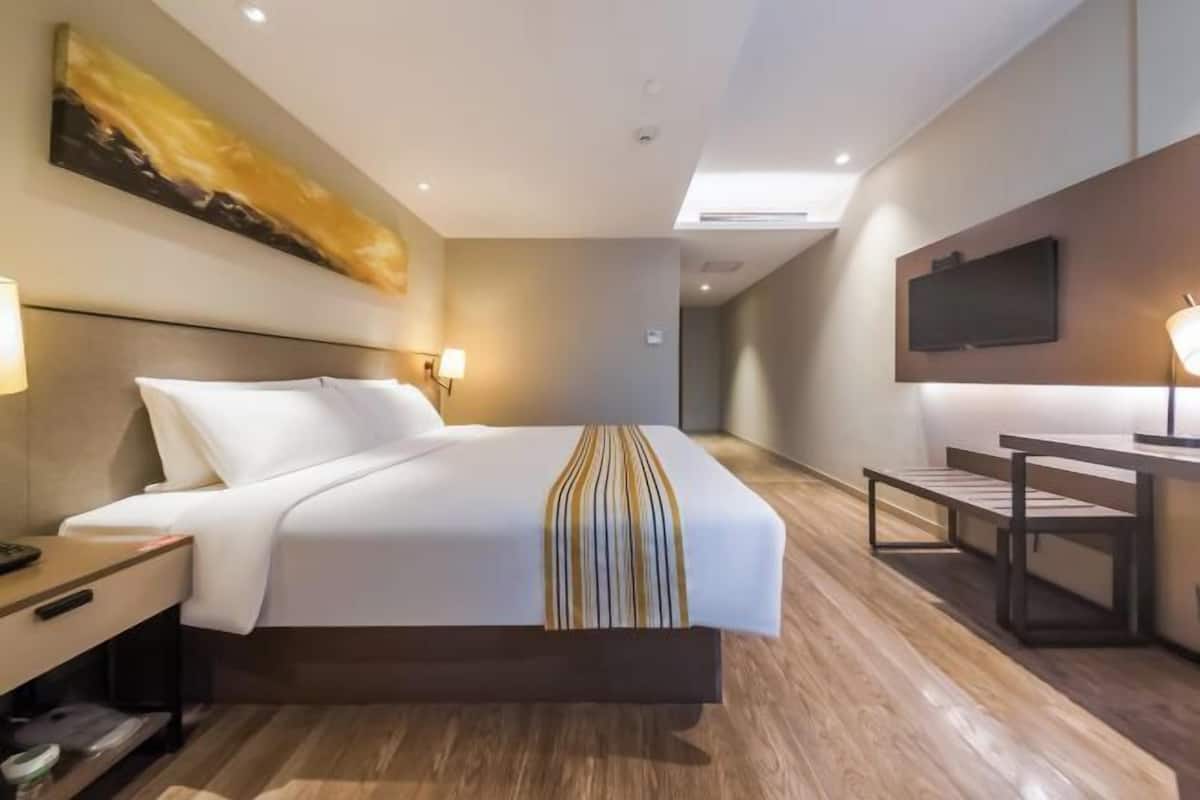 A hotel room with a bed, bedside table, lamp, TV, and a desk with a chair.