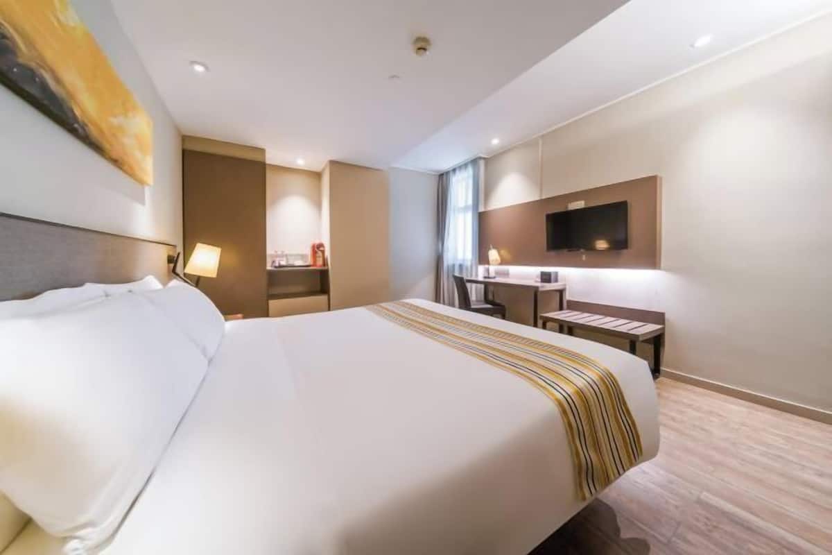 A hotel room with a large bed, a desk, a chair, a flat-screen TV mounted on the wall, and a window with curtains.