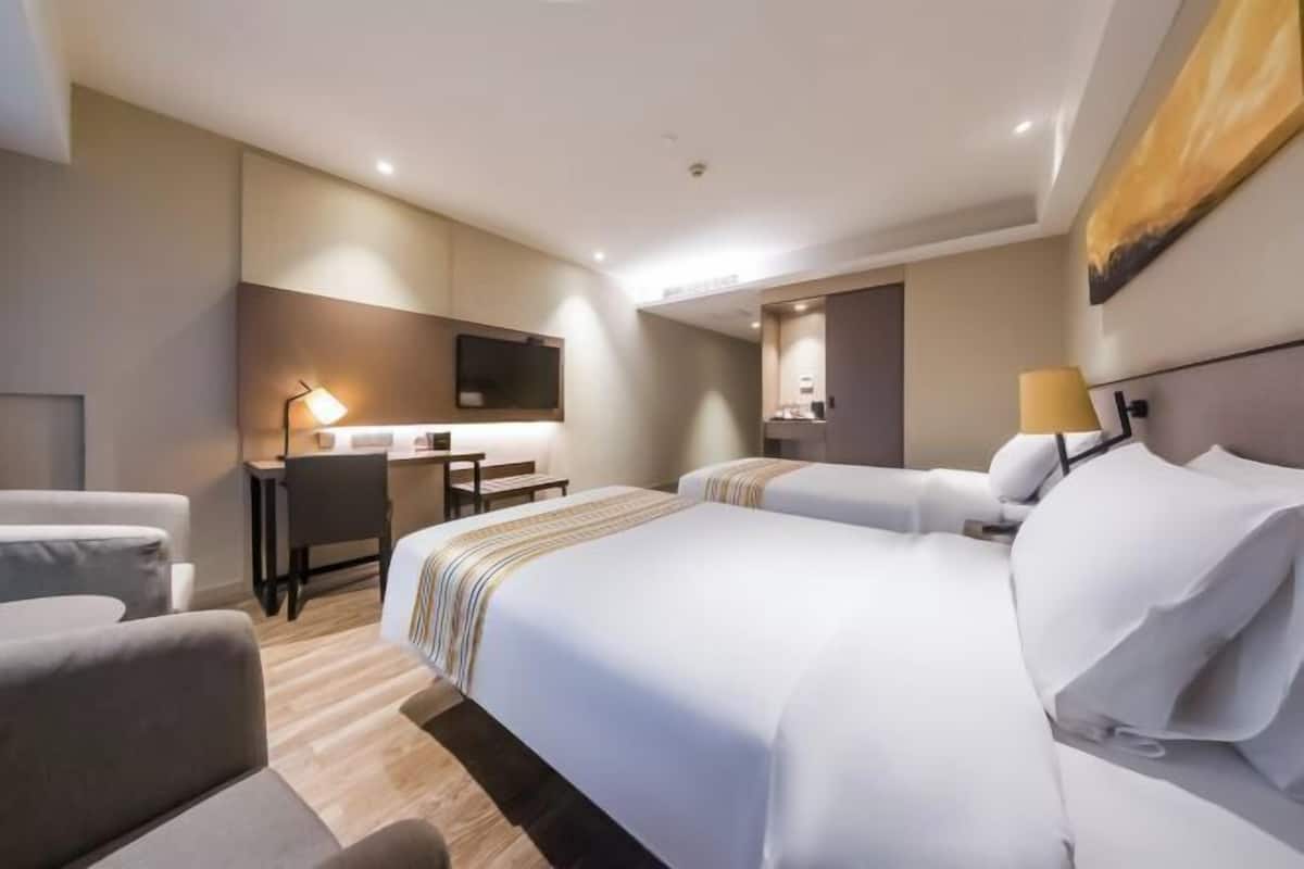 A hotel room with two beds, a desk, a chair, a TV, and a lamp.