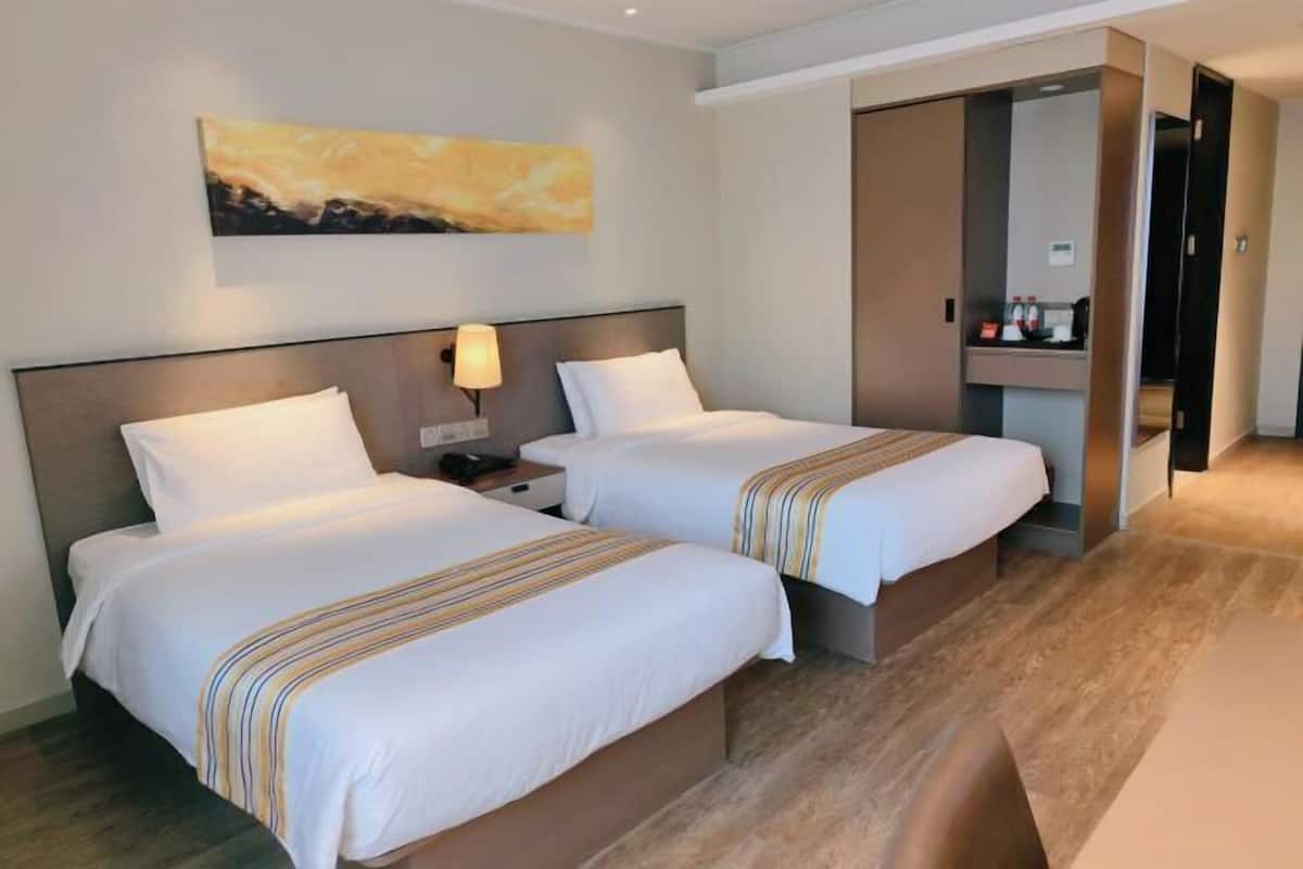 A hotel room with two beds, a nightstand, a lamp, a coffee machine, and a painting on the wall.