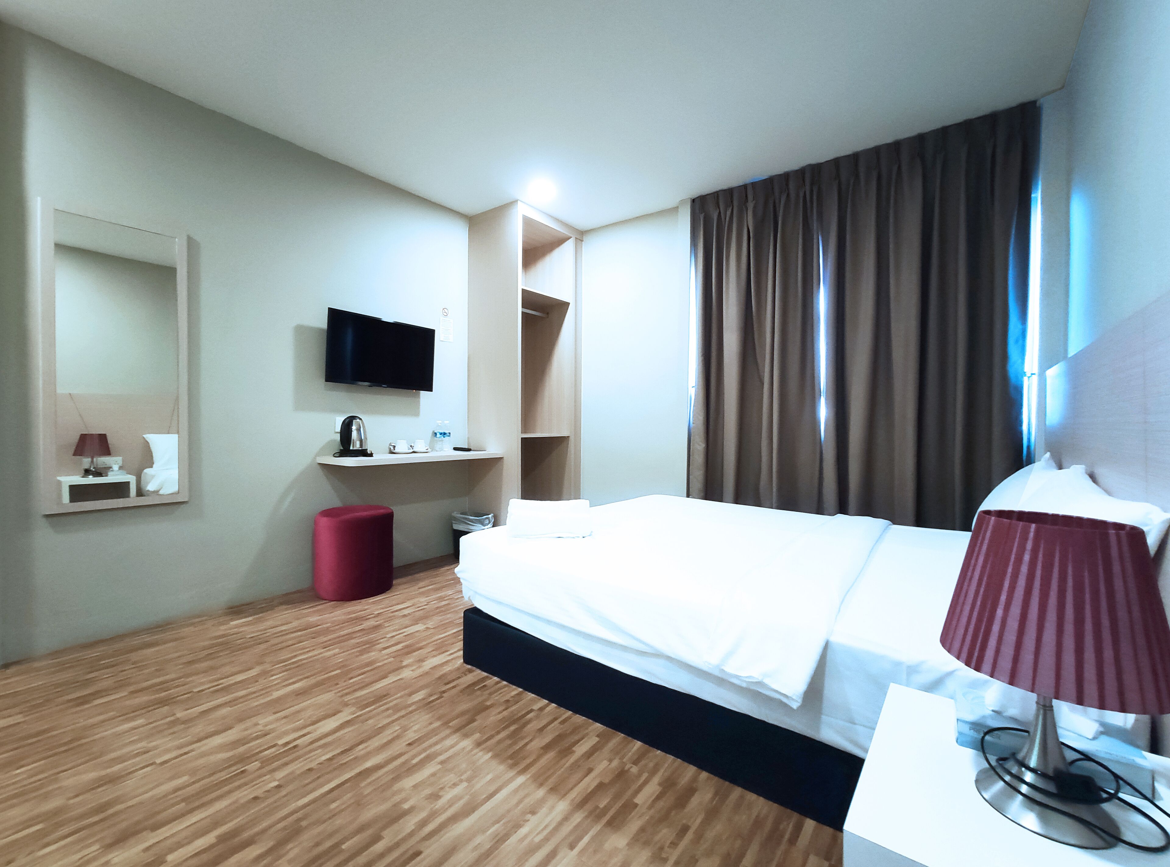 A hotel room with a bed, a TV mounted on the wall, a small table with a lamp, and a red stool.