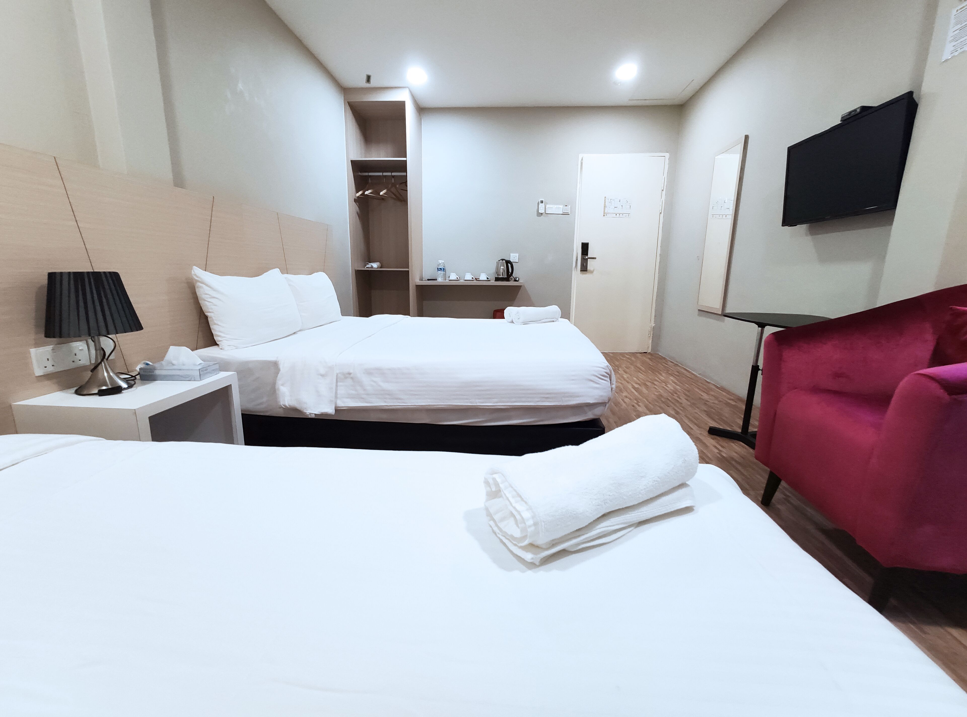 Quarto de hotel com duas camas, um guarda-roupa, uma TV e uma cadeira vermelha.