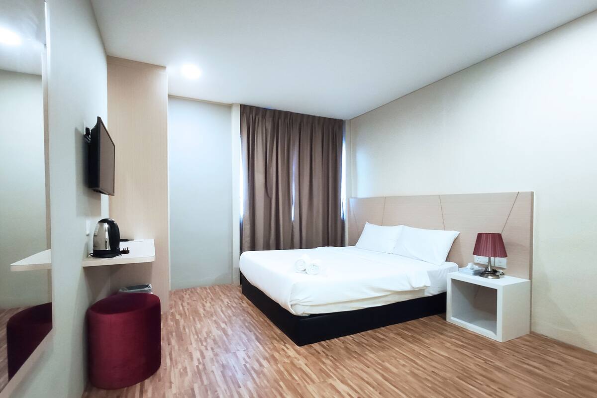 A hotel room with a bed, a bedside table with a lamp, a red ottoman, a wall-mounted TV, and a window with curtains.