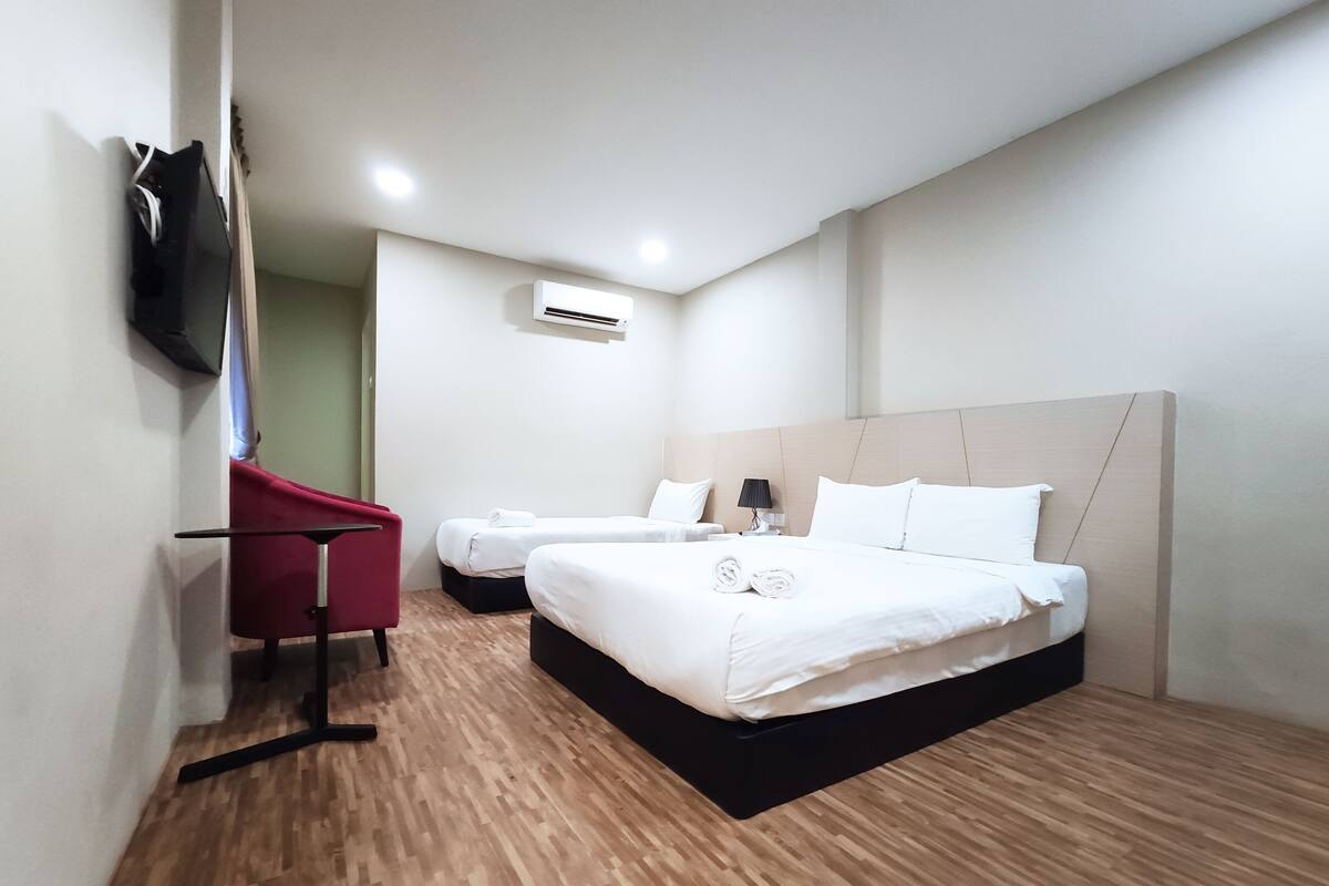 A hotel room with two beds, a red chair, a flat-screen TV, and an air conditioning unit.