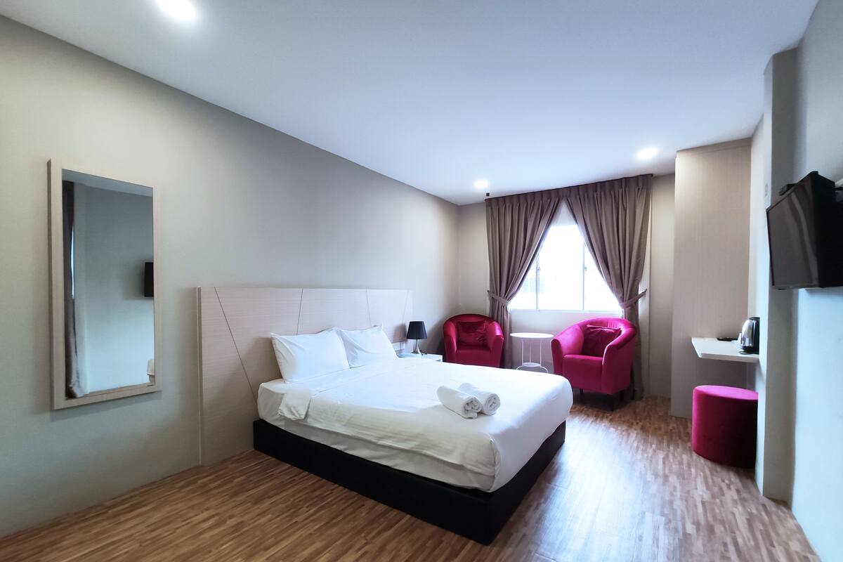 A hotel room with a large bed, two red chairs, a small table, a TV, and a window with curtains.