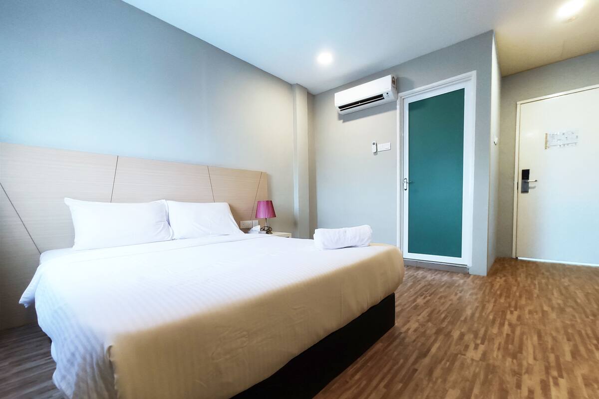 A hotel room with a large bed, a bedside lamp, an air conditioning unit, and a door with a green panel.