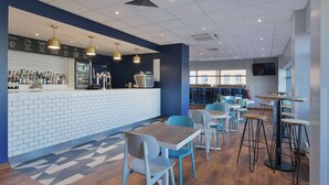 A modern cafe with a bar area, seating arrangements, and a large window providing natural light. - Travelodge Camberley Central (Camberley)