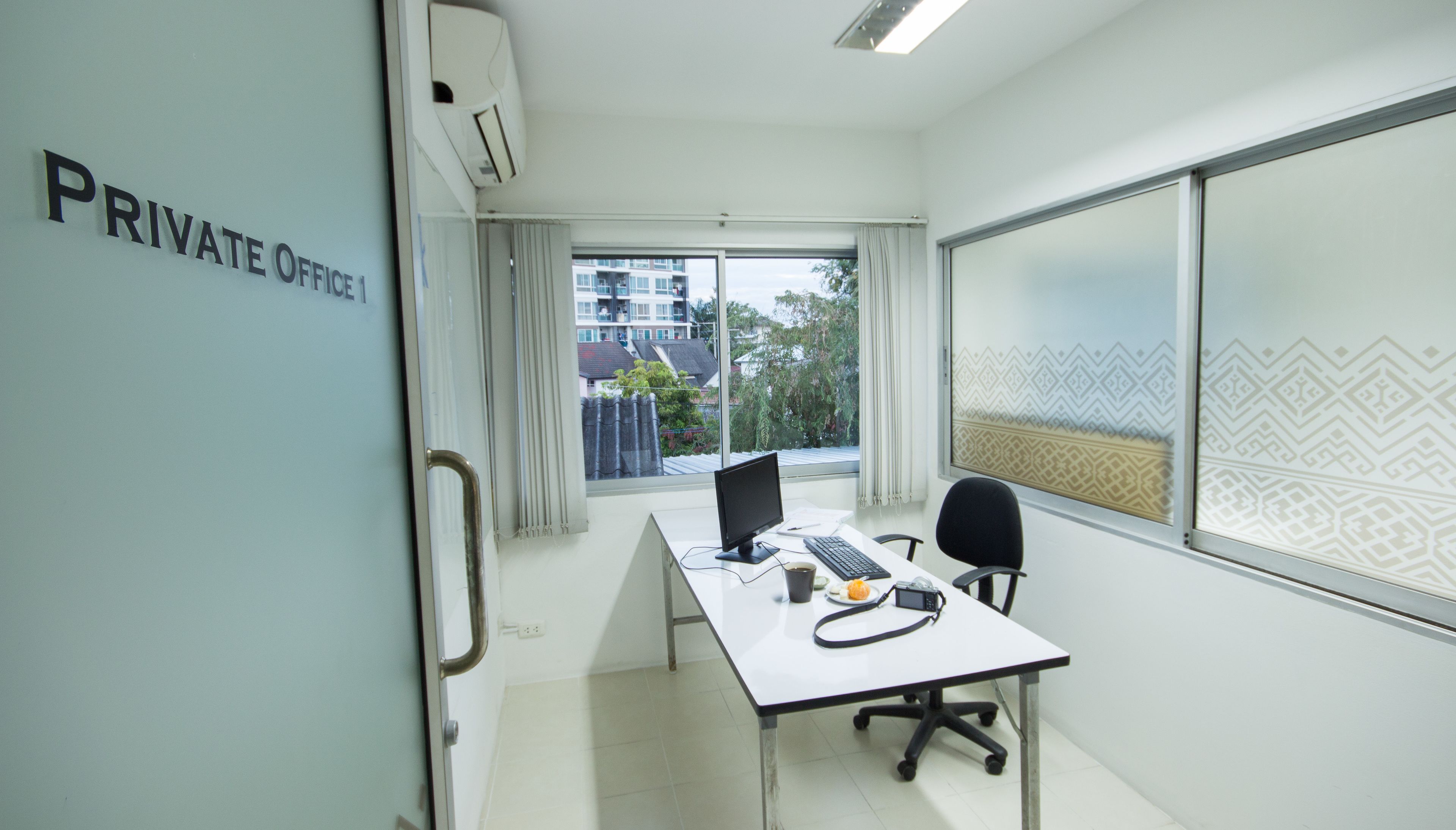 Oficina privada con escritorio, computadora y silla. Hay una ventana que ofrece vistas a edificios y árboles.
