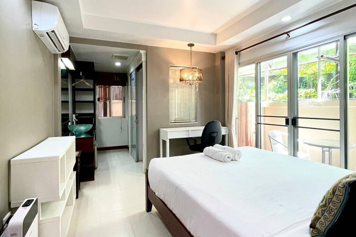 A hotel room with a large bed, a desk, a chair, and a balcony with a view of greenery.