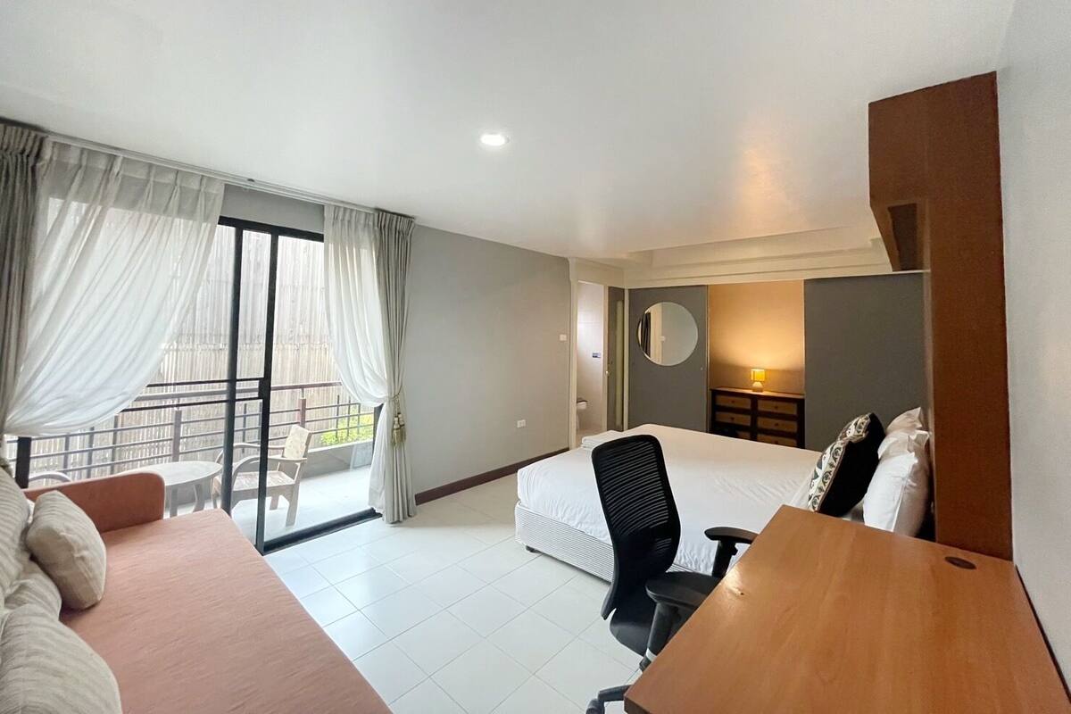 A hotel room with a bed, a desk, a chair, a round mirror, and a balcony with a railing.