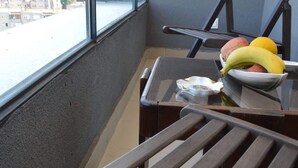 Un balcon donnant sur un paysage urbain, une chaise en bois, une petite table avec un bol de fruits et un mégot de cigarette posé sur la table.