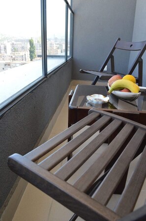 Een balkon met uitzicht op een stadsgezicht, een houten stoel, een kleine tafel met een fruitschaal, en een peuk op de tafel.